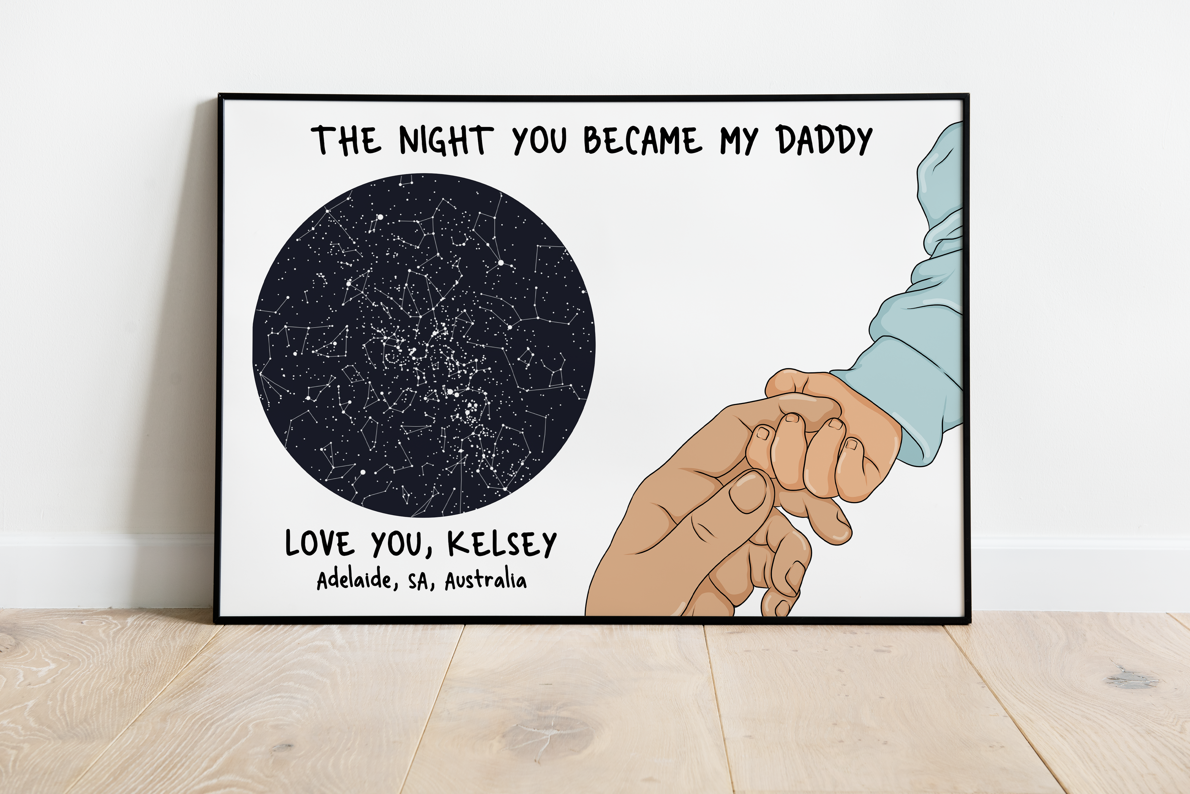 A personalised star map print with the text 'The night you became my daddy' and a blue illustration of a child's hands reaching out to an adult's. There is a wooden frame around the print, placed on a white wall.